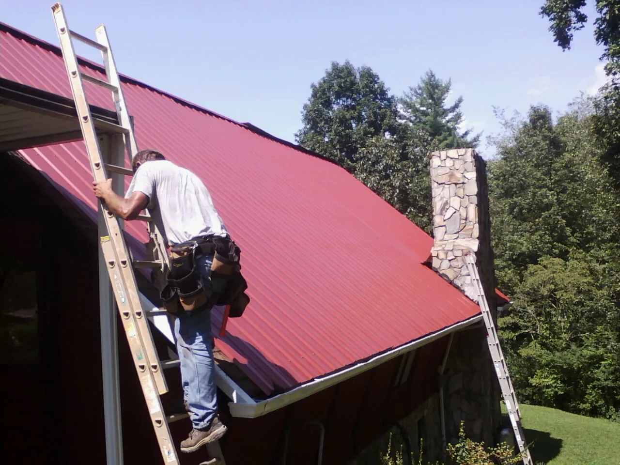 Roofer on a ladder beside a red metal roof and stone chimney — Soffit & Fascia Repair by Roofing Brothers in Haswell